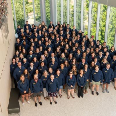 A large group of people in matching navy blue jackets poses 为 a group photo in a bright indoor space with tall windows and greenery outside.